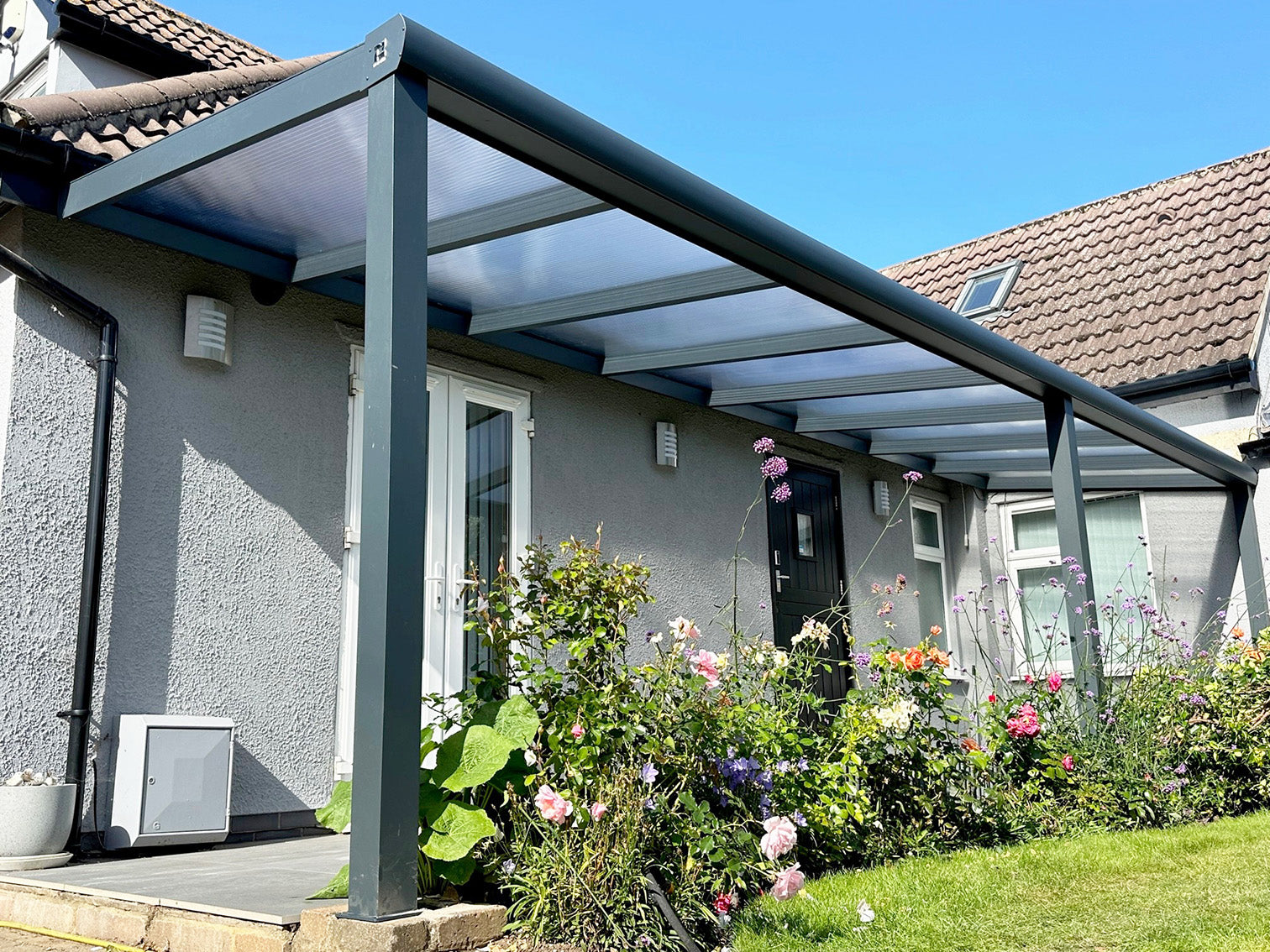 Modern aluminium patio cover with clear roof, attached to house over garden and flowers
