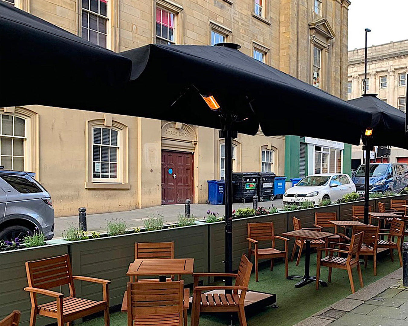 Outdoor dining area with wooden tables, luxury umbrellas, and heaters on a city street