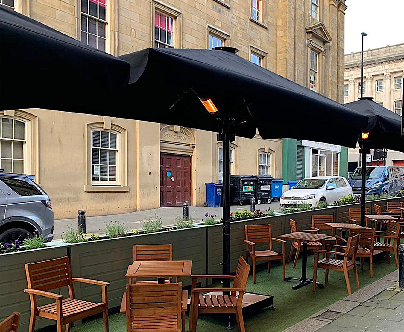 Outdoor dining area with wooden tables, luxury umbrellas, and heaters on a city street