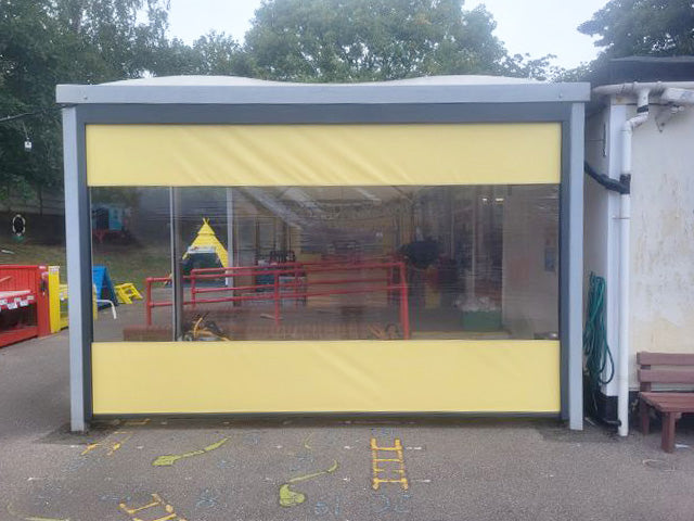 Covered outdoor structure with yellow retractable screens and clear panel, ideal for playground or patio.